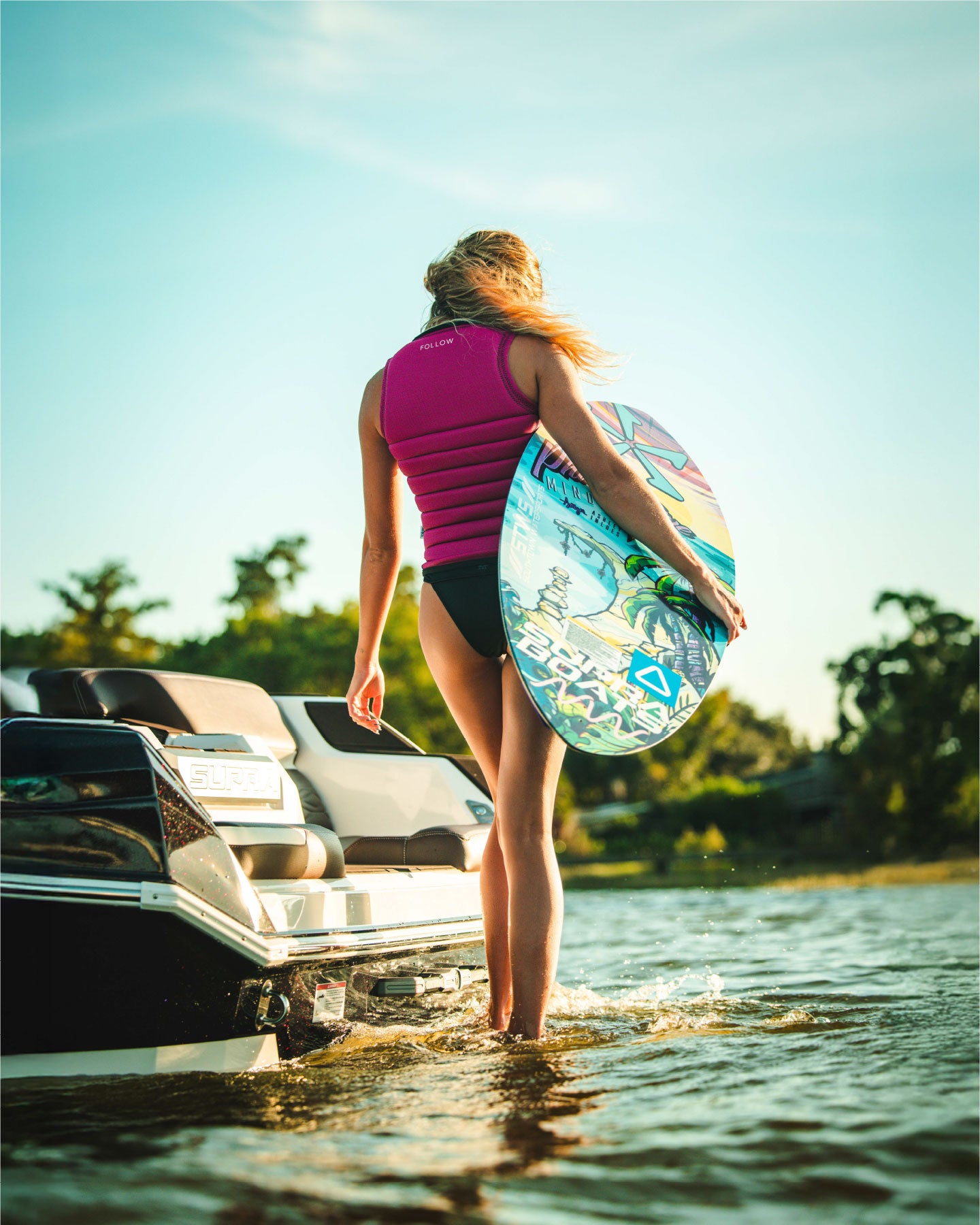 Person holding a colorful surfboard near a boat on a sunny day