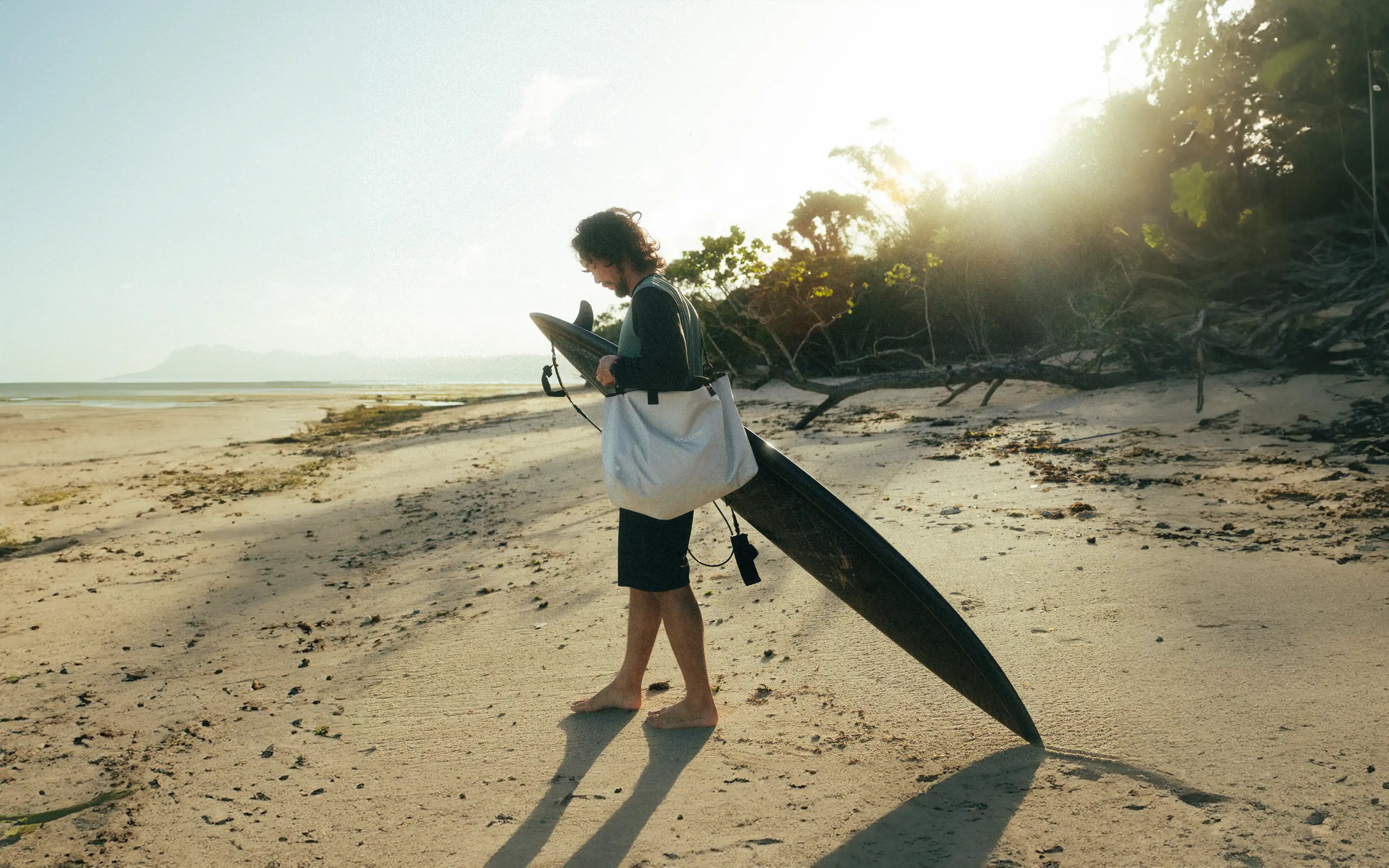Josh Ku holding a surfboard on a beach with the sun shining through trees