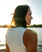 Woman wearing a white tank top with 'FOLLOW' text by a body of water.