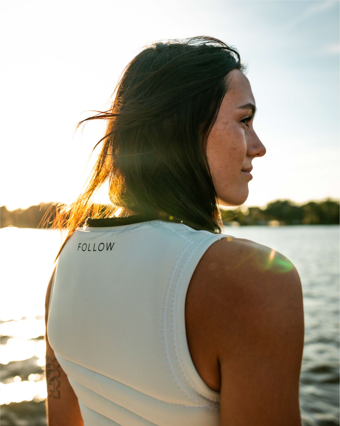 Woman wearing a white tank top with 'FOLLOW' text by a body of water.